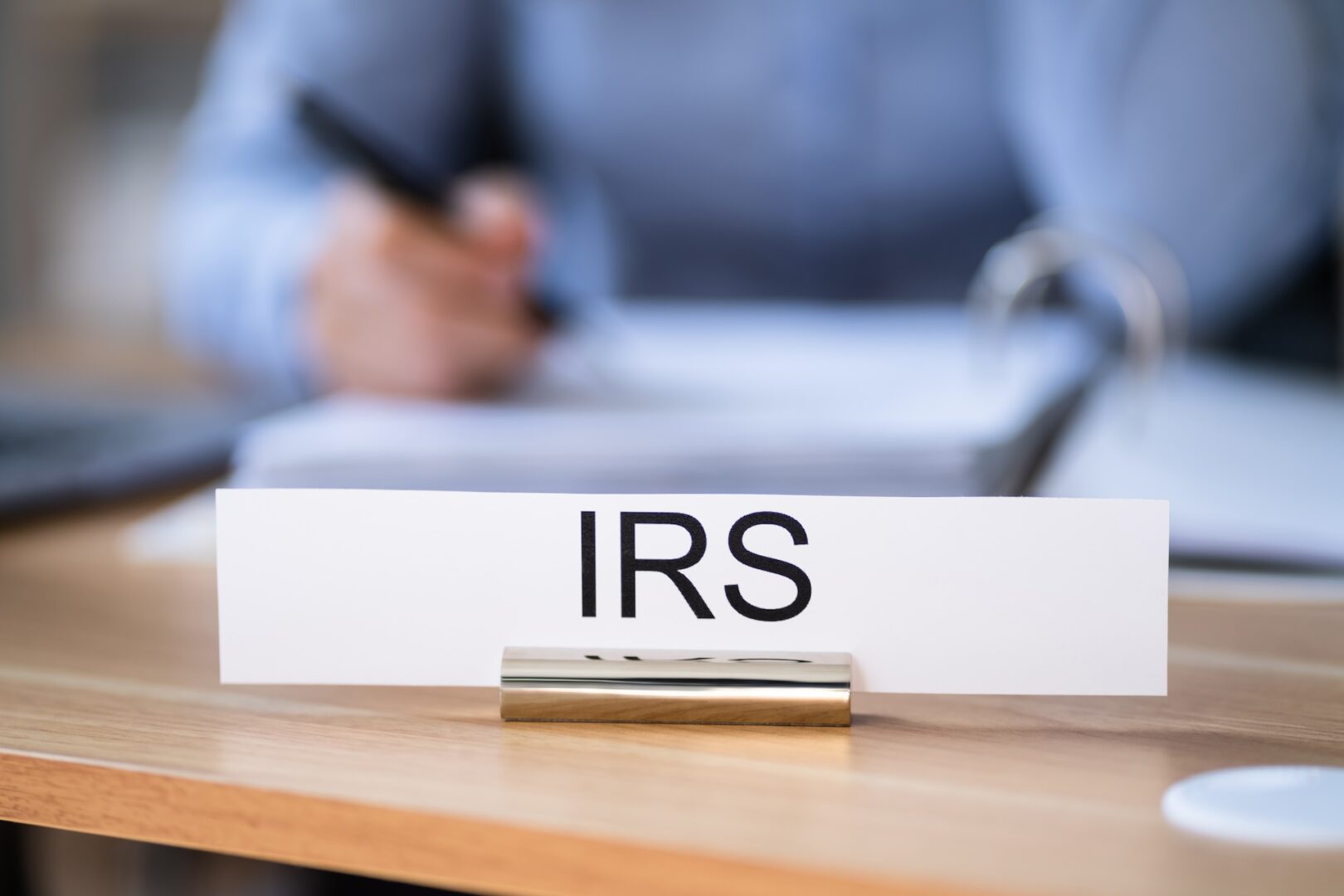 A nameplate with "IRS" written on it is displayed on a desk. In the background, a person is blurred, seemingly working with documents, highlighting a setting reminiscent of a governmental office or official work environment.