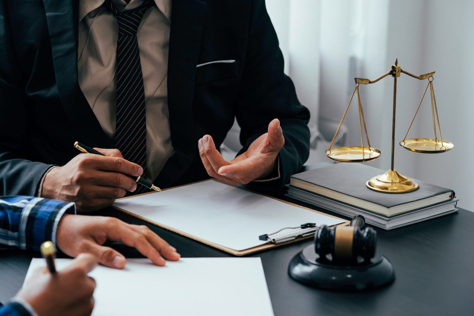 Two people are seen discussing legal documents at a desk. One person is holding a clipboard and pen, gesturing with their hand, while the other is writing notes. A pair of scales, a stack of books, and a pair of headphones are also on the desk.