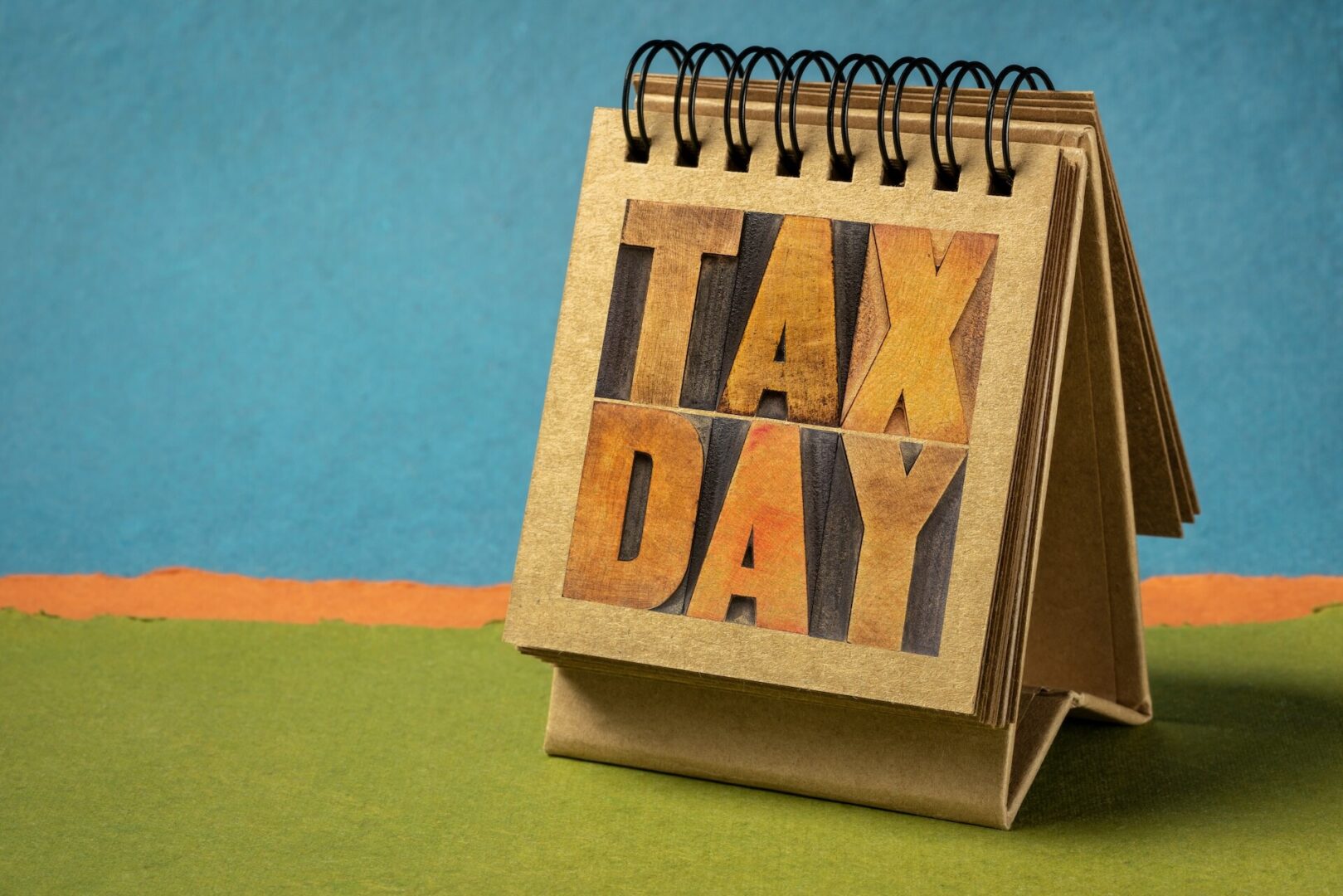 A small desktop calendar with a spiral binding displays the words "TAX DAY" in large, bold letters. The background is a mix of blue, orange, and green paper, giving a textured and colorful backdrop to the calendar.