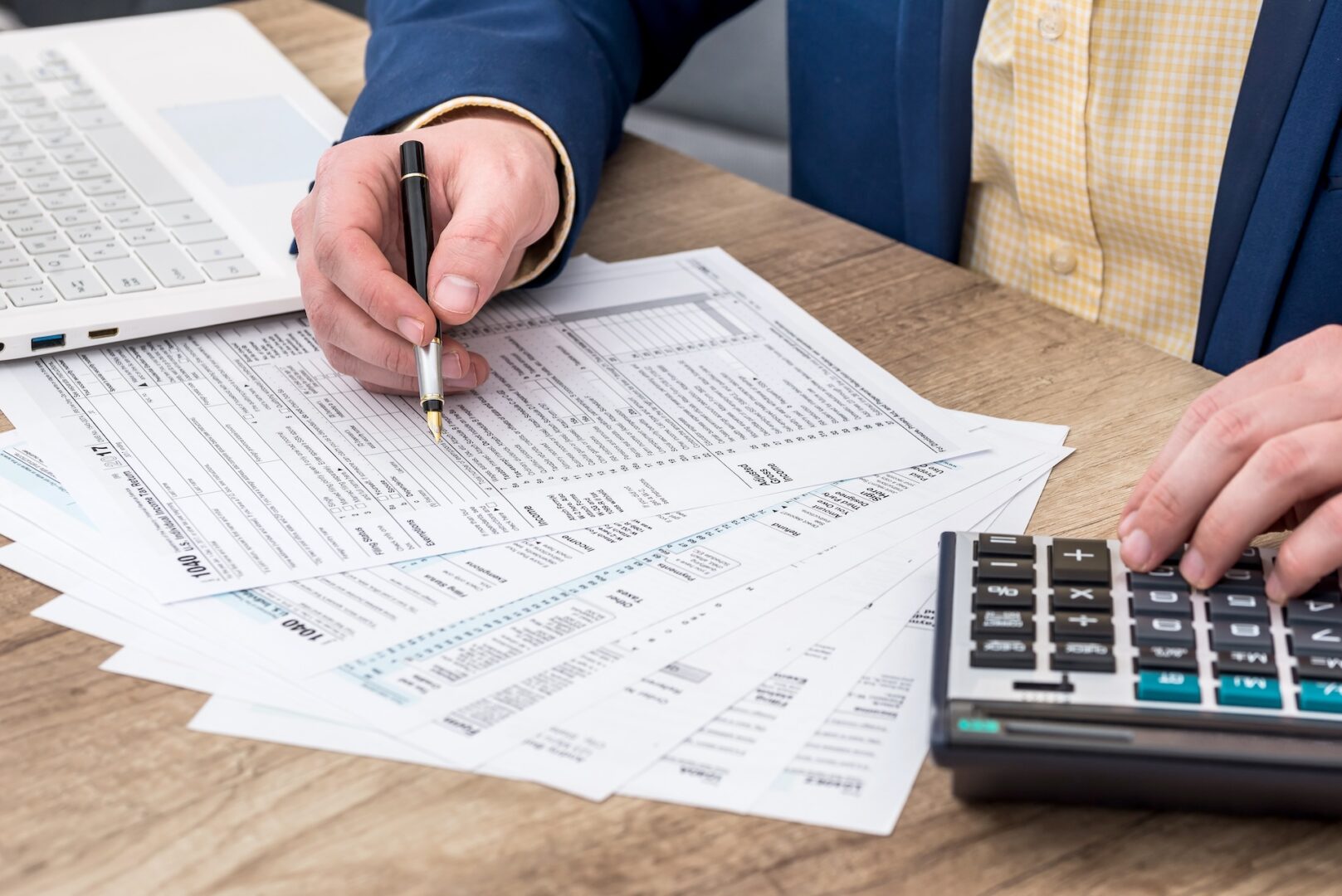 A person in a blue blazer reviews financial documents at a desk. One hand holds a pen over the papers, while the other hand types on a calculator. A laptop is open on the left side of the desk. The setting appears to be an office or a workspace.