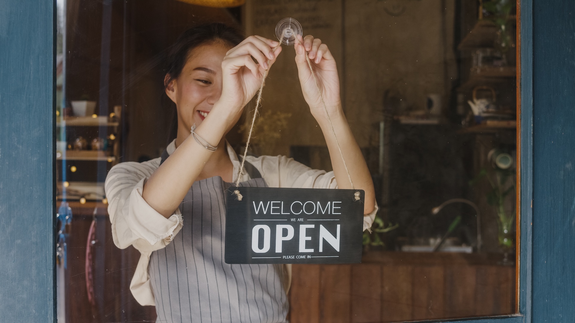 A person wearing a gray apron is smiling and hanging an "Open" sign on the glass door of a shop or café. The sign reads "Welcome, we are OPEN." The background shows a cozy interior with wooden elements and plants.