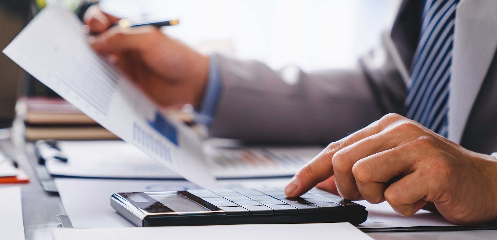 A person wearing a suit is holding a document in one hand and using the other hand to press buttons on a calculator. The scene is set on a desk with papers and a pen, suggesting a business or financial setting. A person wearing a suit is holding a document in one hand and using the other hand to press buttons on a calculator. The scene is set on a desk with papers and a pen, suggesting a business or financial setting.