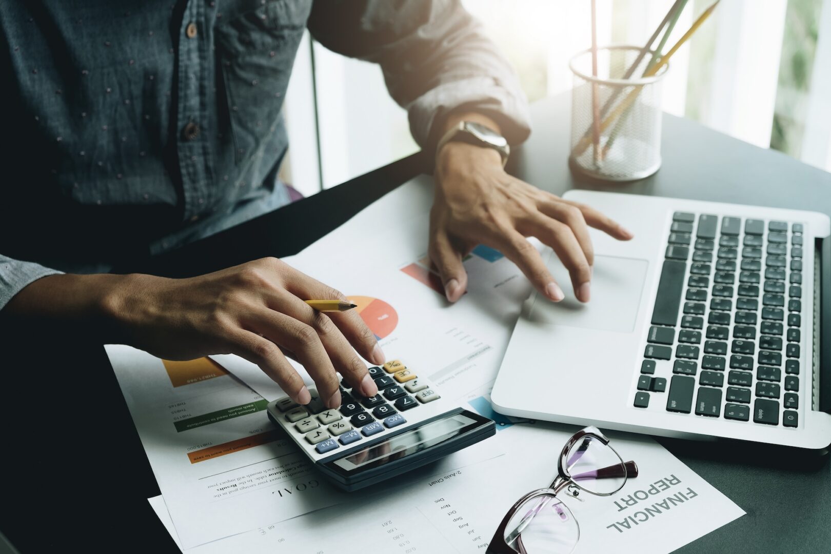 A person uses a calculator and a laptop at a desk cluttered with financial charts and documents. Nearby are a pair of glasses and a pen inside a holder. Sunlight streams in from a window.