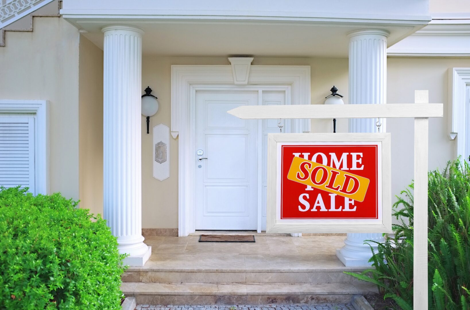 A house with a white front door and columns is shown. A red sign with white and yellow text reading "Home Sold Sale" is displayed on a post in the front yard, surrounded by green shrubbery.