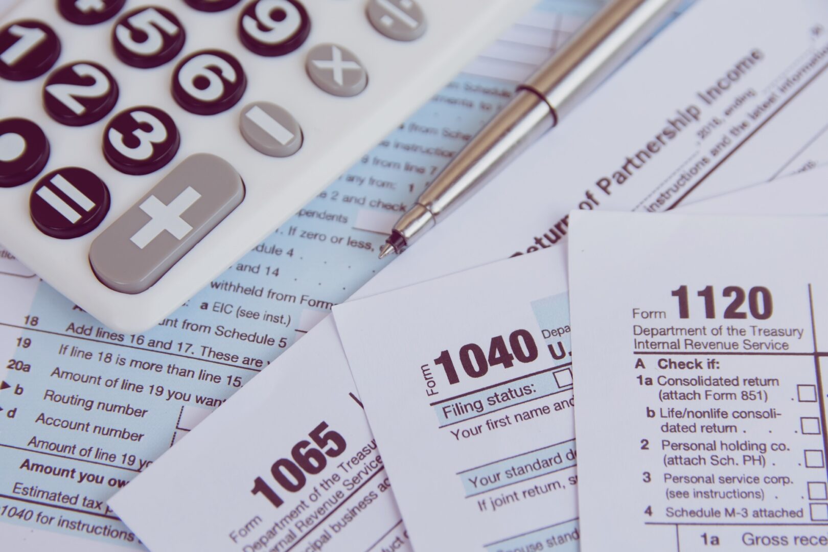 A calculator and a pen rest on a pile of U.S. tax forms, including 1040, 1065, and 1120. The forms are partially visible, suggesting a tax preparation or financial planning scene.