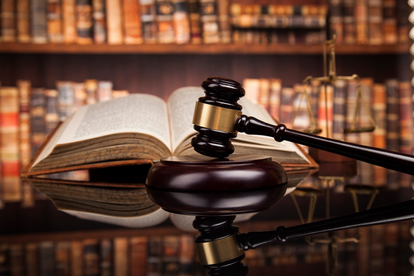 A wooden gavel rests on a sound block in the foreground, with an open book and a scale of justice in the background. The setting is a library or study with shelves filled with books.