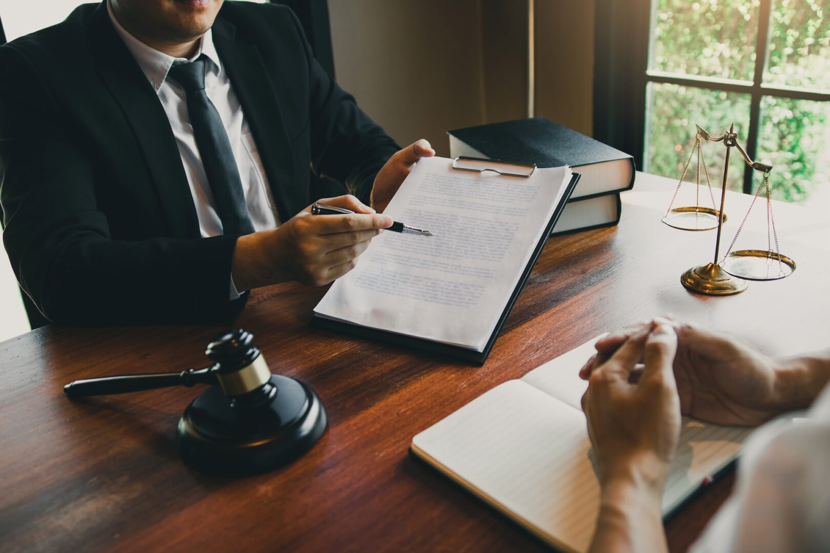 Two people are seated at a wooden desk discussing legal documents. One person holds a clipboard with papers, using a pen for emphasis. A gavel, scales of justice, and stacked books are on the table, suggesting a legal or courtroom setting.