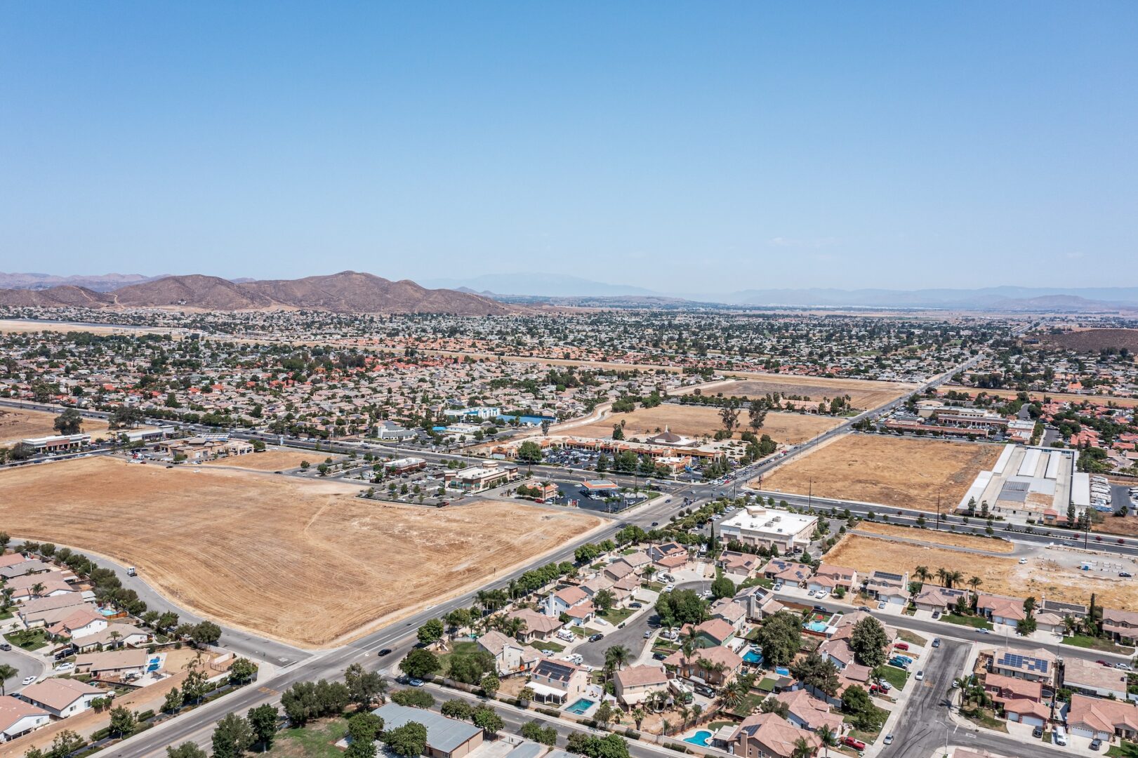 Aerial view of a suburban area with a grid layout. There are houses, roads, and open fields. The landscape is flat with distant mountains under a clear blue sky.