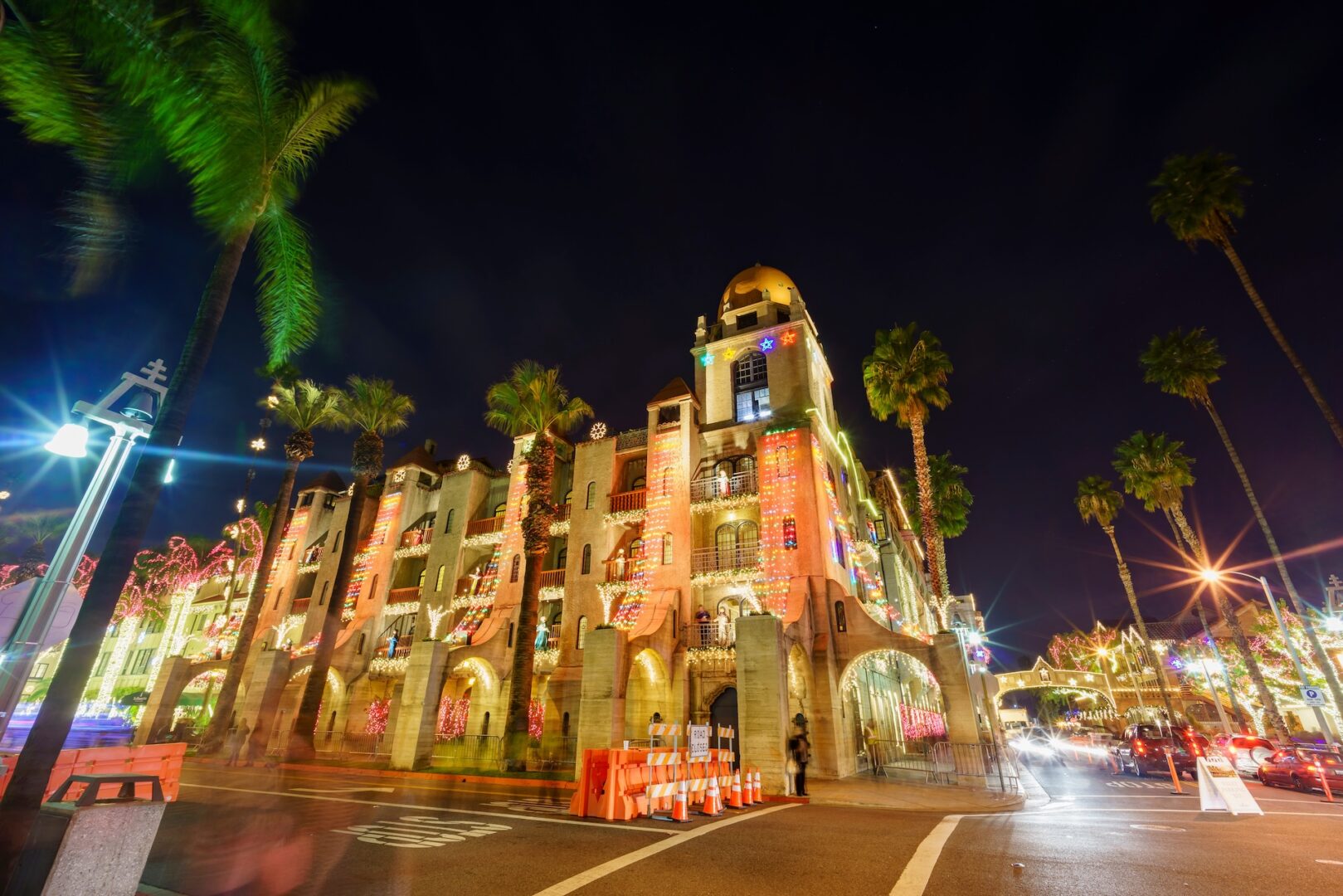 A festive building illuminated with colorful lights at night, surrounded by palm trees. Traffic cones and blurred car lights are visible in the foreground, adding a sense of motion to the scene.