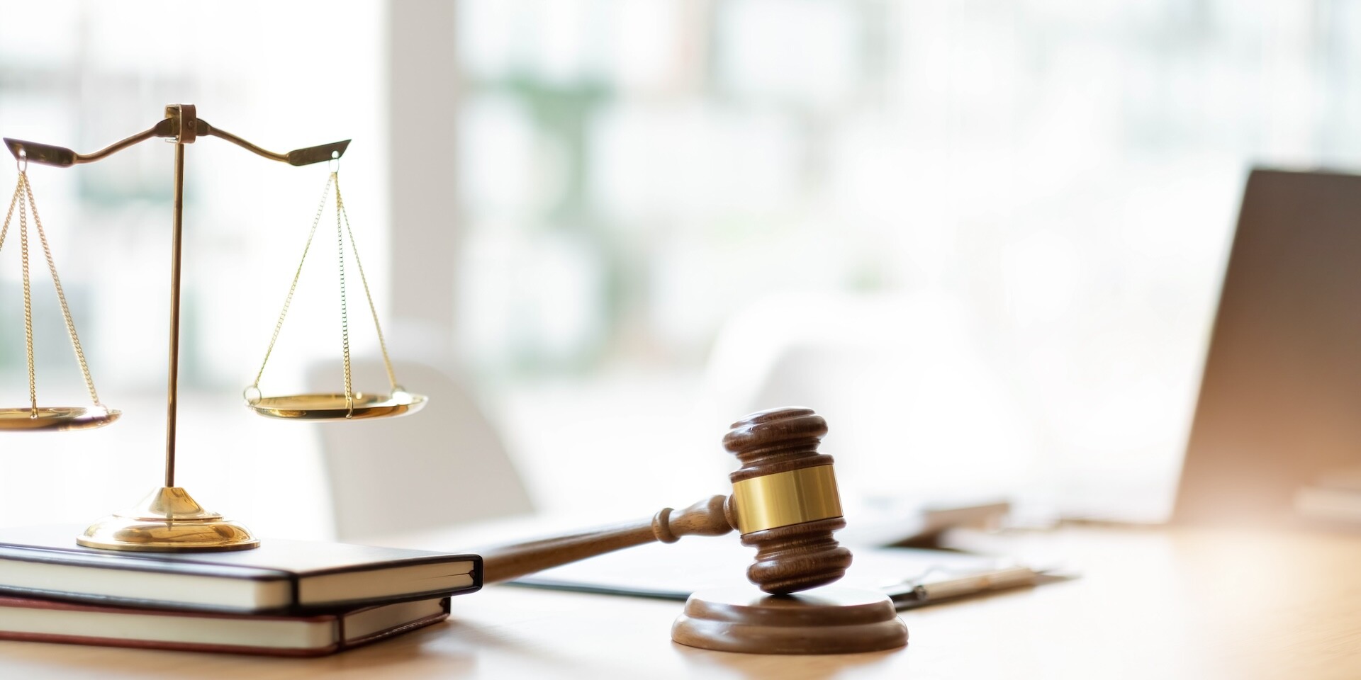 A close-up of a wooden judge's gavel and a balanced scale on a desk, with stacked books and a blurred background. The scene conveys a legal or judicial setting, emphasizing justice and law.