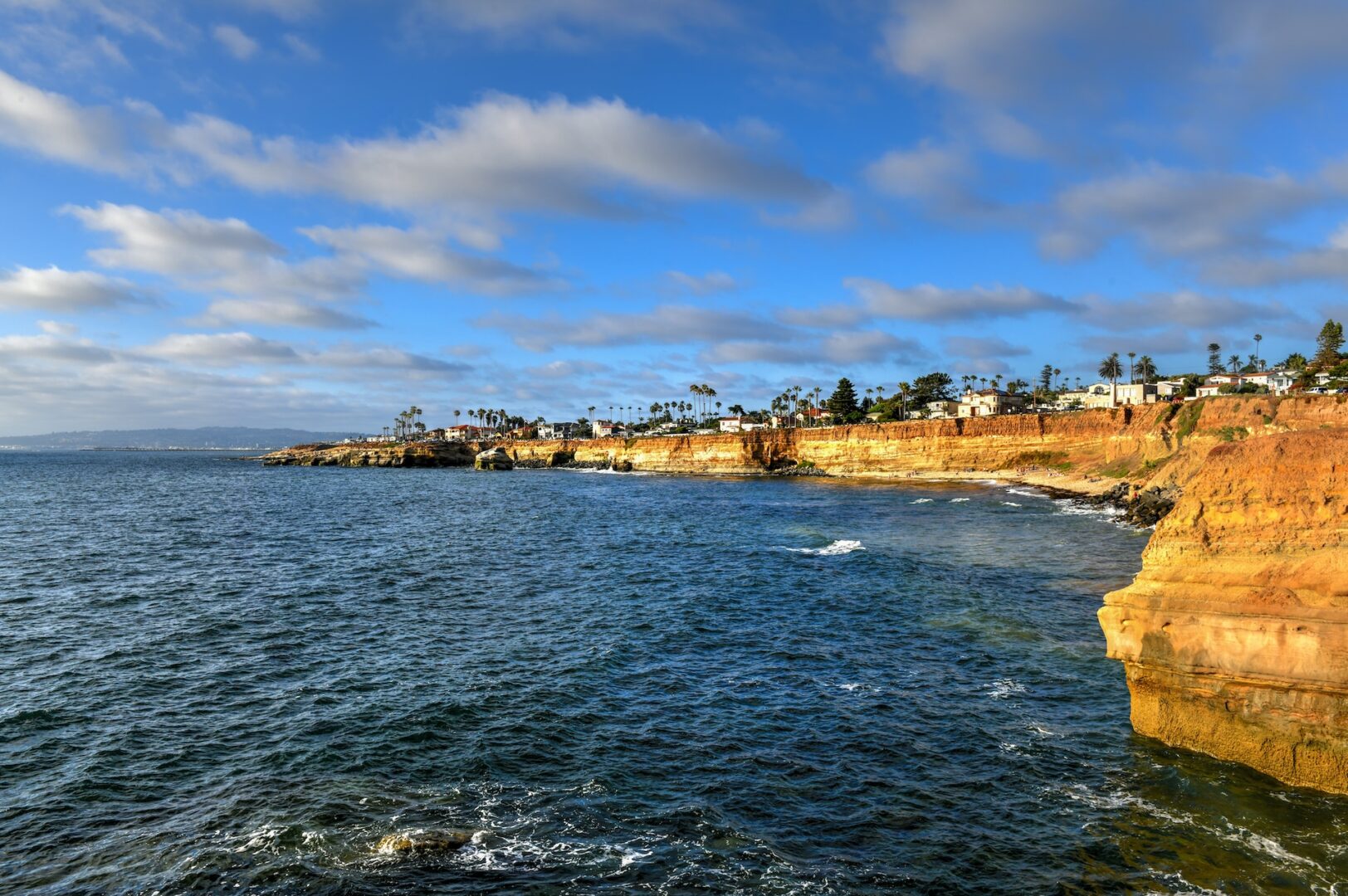 Coastal view of rocky cliffs meeting the ocean under a partly cloudy sky. The shoreline is lined with palm trees and buildings, creating a picturesque scene of nature and human habitation. The water is calm with gentle waves.