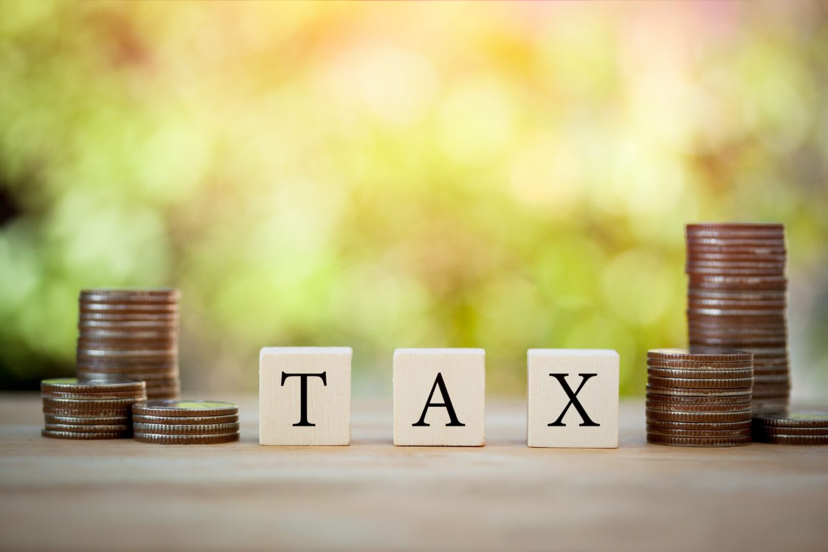 Stacks of coins are placed on either side of wooden blocks spelling "TAX" on a wooden table. The background is softly blurred with shades of green and yellow, suggesting an outdoor setting.