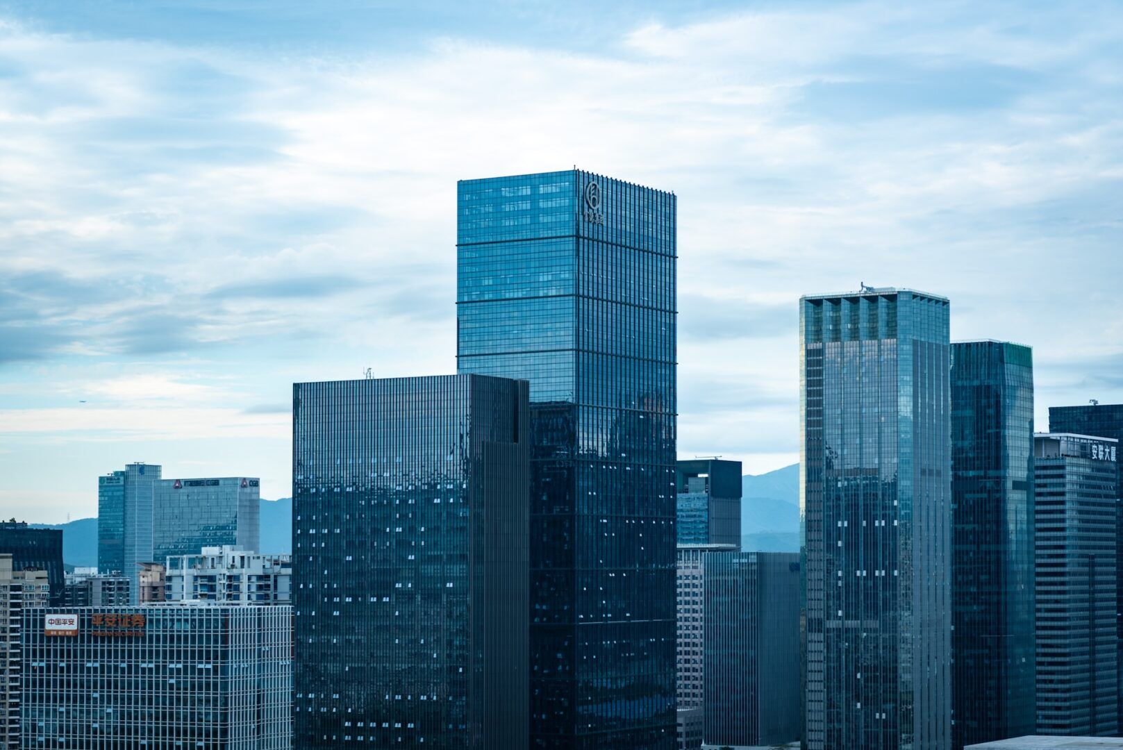 City skyline with modern glass skyscrapers under a partly cloudy sky. The tallest building is centrally located, reflecting the blue sky and clouds. Smaller buildings surround it, creating a dense urban landscape.