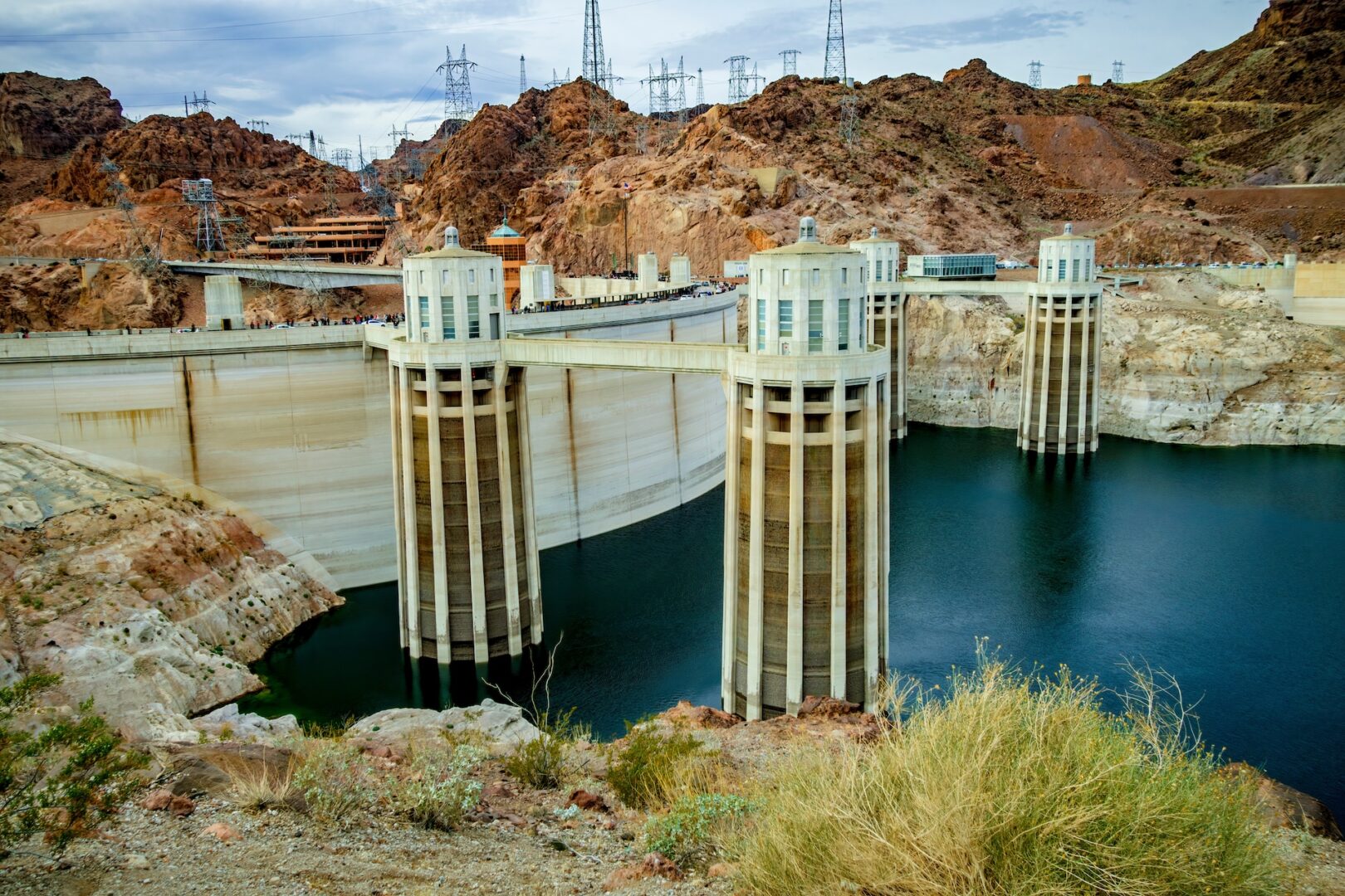 A large dam with tall water intake towers spans across a deep blue reservoir, surrounded by rugged, reddish-brown rocky terrain. Power lines stretch across the background under a cloudy sky. Sparse vegetation is in the foreground.