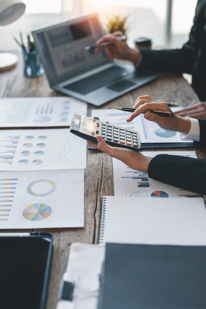 Close-up of two people in business attire at a wooden table with a laptop, holding pens and a calculator. The table is covered with financial charts and graphs, papers, and a notebook, suggesting a meeting or financial analysis session.