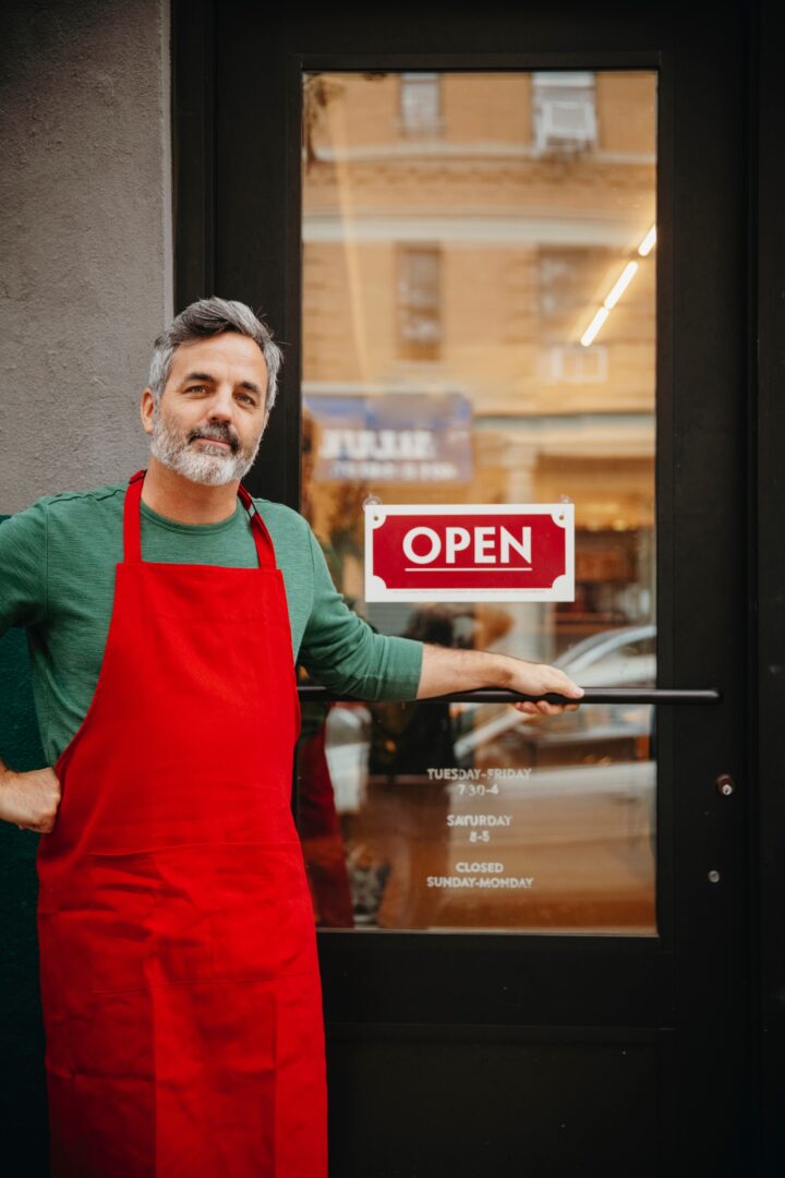 A man in a red apron stands smiling at the entrance of a shop with a glass door. The door displays an "OPEN" sign and the store hours. The background shows a blurred street view.