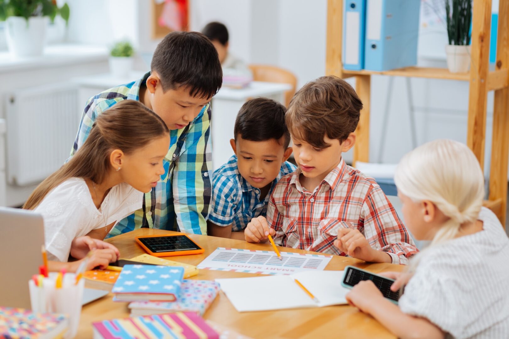 Five children are gathered around a table in a classroom, focused on a group activity. They are looking at papers and holding pencils and calculators. The table is cluttered with school supplies, including notebooks and colored pencils.