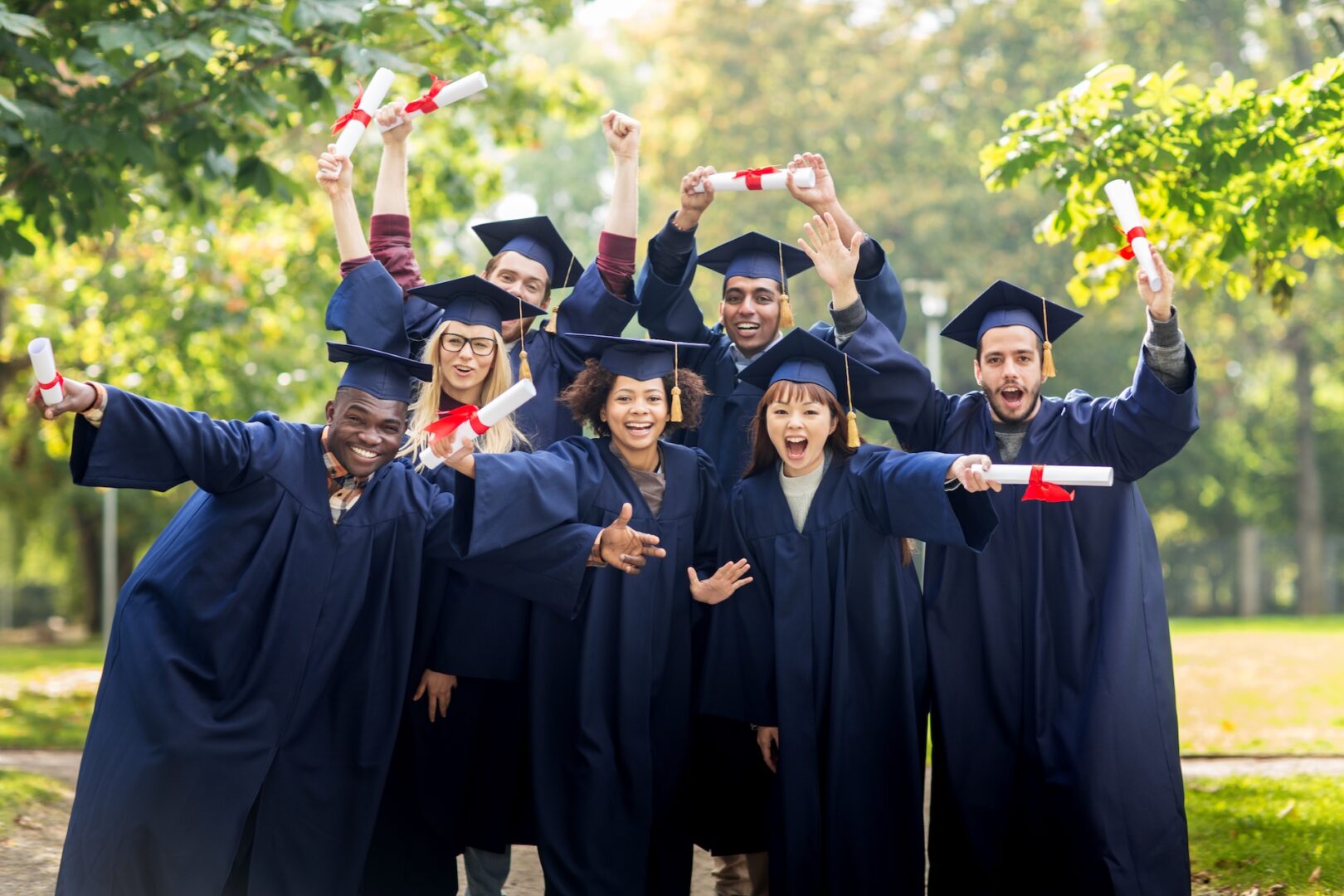 A group of seven graduates in blue gowns and caps celebrate outdoors, holding diplomas and cheering. They appear joyful, surrounded by green trees in a park setting.