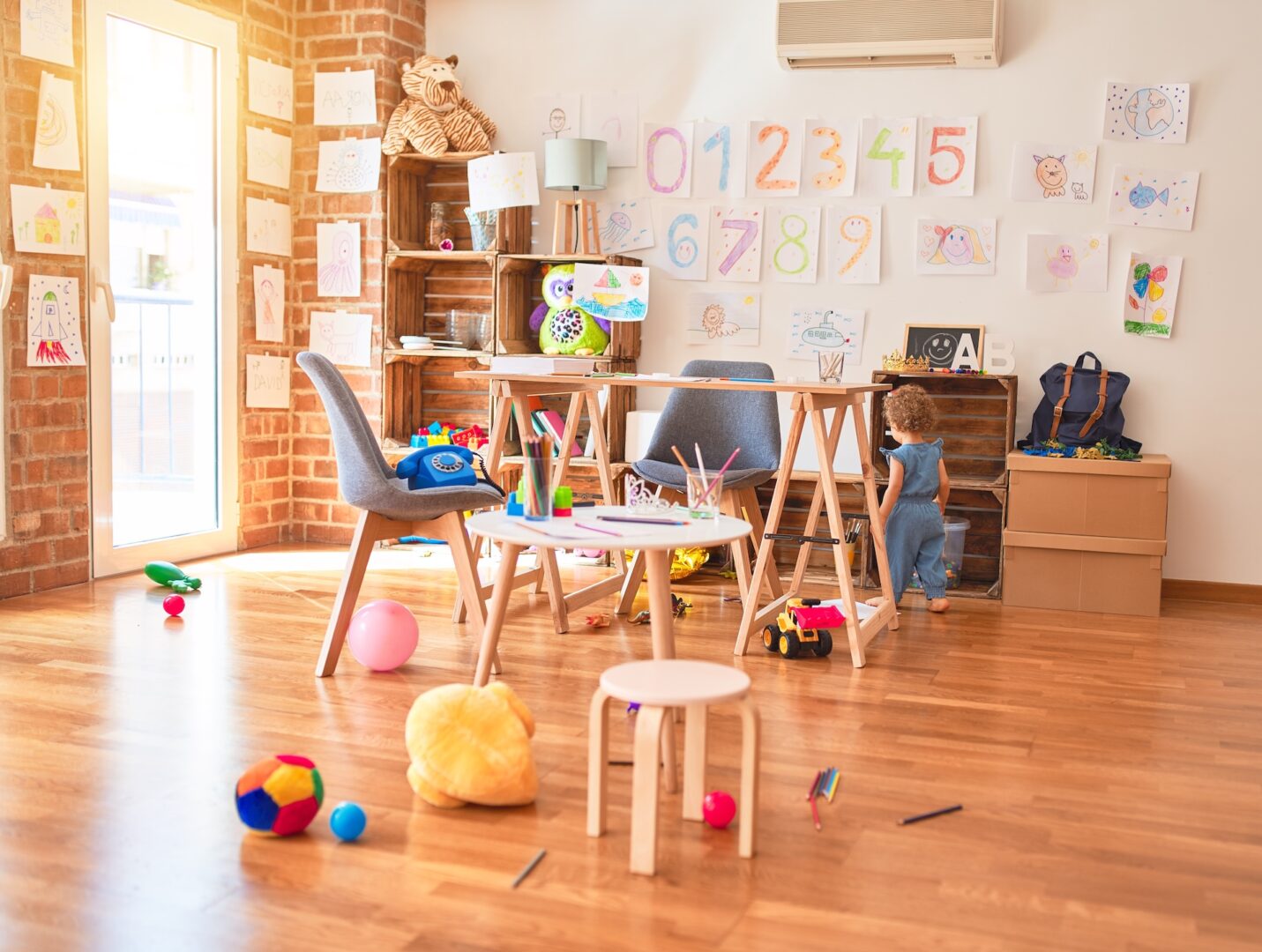 A child in a blue outfit stands in a bright playroom with wooden floors. The room is scattered with colorful balls and toys. Drawings and numbers decorate the walls. Two small tables and several chairs complete the cheerful, creative space.