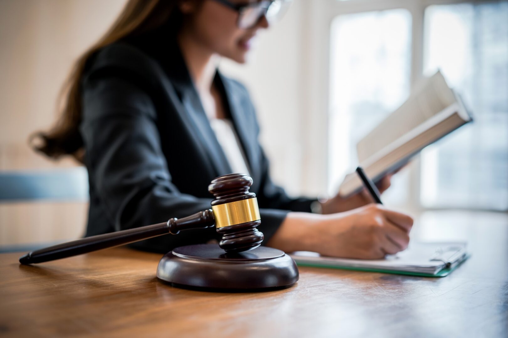 A judge’s gavel rests on a desk in the foreground, while a woman in business attire sits in the background, writing notes in an open book. The setting appears to be a legal office or courtroom. A judge’s gavel rests on a desk in the foreground, while a woman in business attire sits in the background, writing notes in an open book. The setting appears to be a legal office or courtroom.