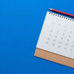 A spiral-bound desk calendar showing a month, placed on a brown envelope, with a red pencil above it on a bright blue background—a useful setup for A Guide To Business Formation and Quarterly Taxes.