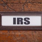 A metal label holder with a white card inside displays the bold black letters "IRS" against a wooden background, hinting at secrets like “10 Tax Tips From a Former IRS Insider” amid the visible grain and cracks.