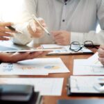 Three people sit at a table in a business meeting, discussing graphs and charts on paper and a tablet. As they review documents about What Are Tax Amendments and How Do I Make Them?, hands gesture with pens while glasses and a laptop rest on the table.