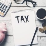 A notepad with the word "TAX" sits on a desk alongside a calculator, glasses, a pink piggy bank, coins, and coffee—perfect for learning about the most popular types of trusts and how to form one.