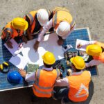 Six construction workers in safety vests and helmets gather around a table with blueprints and electronic components, discussing a project—much like careful estate planning with Will, Trust, or Inheritance? Estate Planning Tips for Californians.