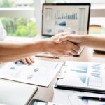 Two people shaking hands over a desk covered with financial charts, documents, and a laptop, reflecting a successful meeting on What Savings Strategies Are Tax Deductible? Guidance from a California Tax Attorney in a bright office setting.