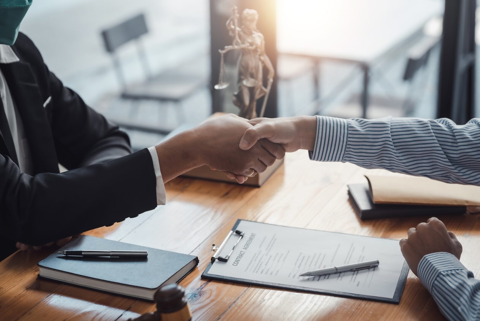 Two people in business attire shake hands across a table with documents, notebooks, and a gavel, suggesting a legal or business meeting in an office setting. Two people in business attire shake hands across a table with documents, notebooks, and a gavel, suggesting a legal or business meeting in an office setting.