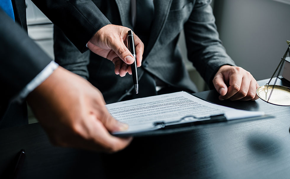 Two people in business suits review and discuss a document on a clipboard at a desk, with one person holding a pen and pointing at the paper. Two people in business suits review and discuss a document on a clipboard at a desk, with one person holding a pen and pointing at the paper.