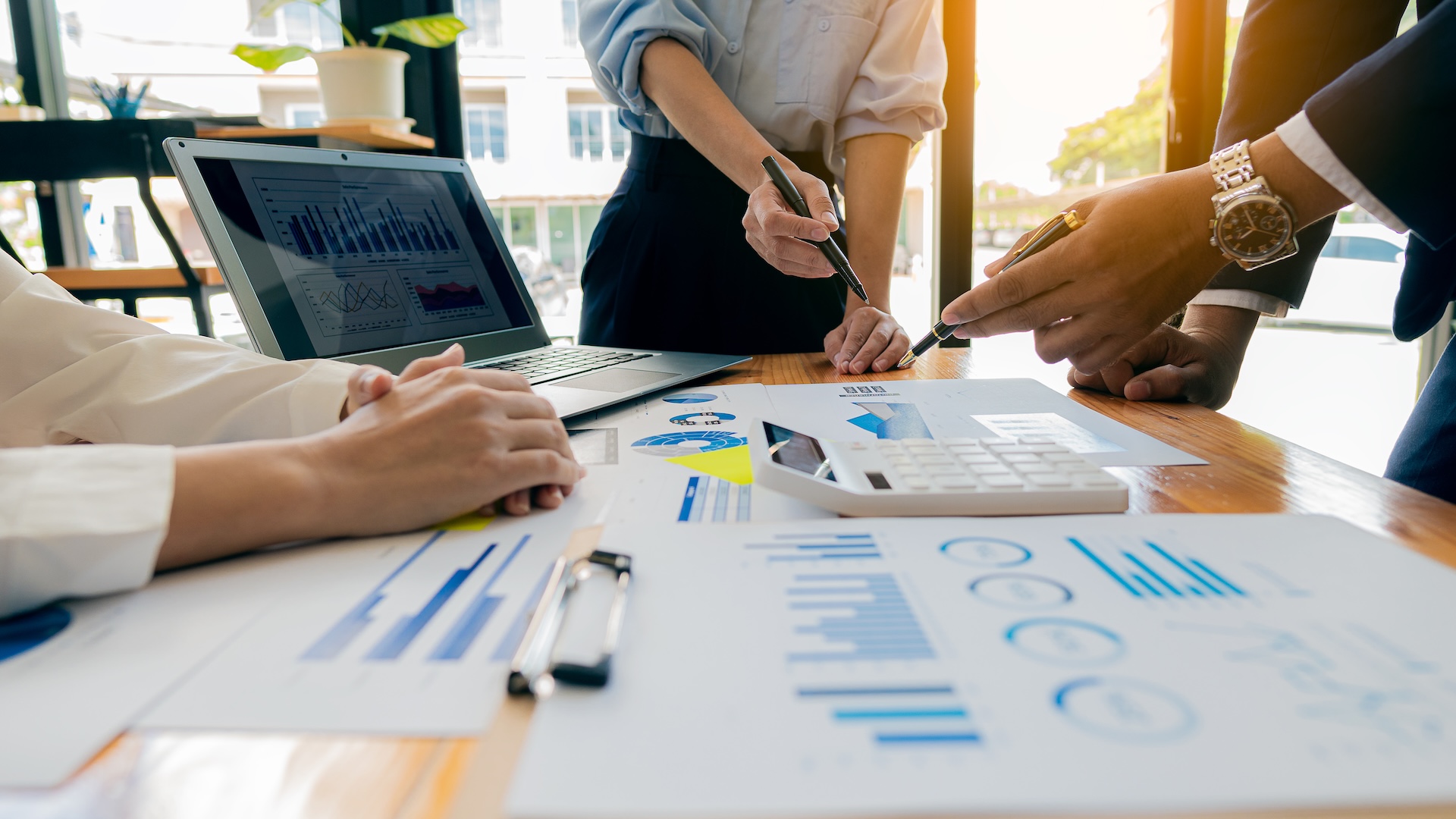 Three people stand around a table analyzing financial charts, graphs, and data reports. A laptop, calculator, pens, and papers are visible, suggesting a business meeting or teamwork on financial planning. Three people stand around a table analyzing financial charts, graphs, and data reports. A laptop, calculator, pens, and papers are visible, suggesting a business meeting or teamwork on financial planning.