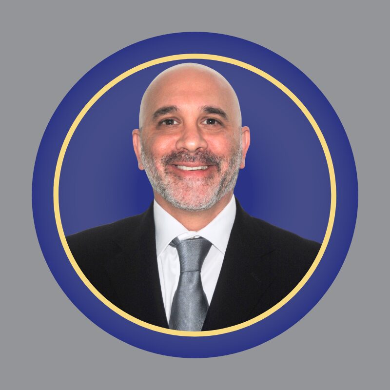 A smiling man with a shaved head and beard, wearing a dark suit, white shirt, and gray tie, is centered in a circular blue and gold border on a gray background.
