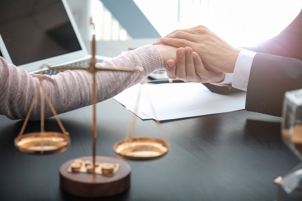 A person in a suit holds another person's hand in a comforting gesture at a desk with legal scales and documents, suggesting a supportive legal consultation. A person in a suit holds another person's hand in a comforting gesture at a desk with legal scales and documents, suggesting a supportive legal consultation.