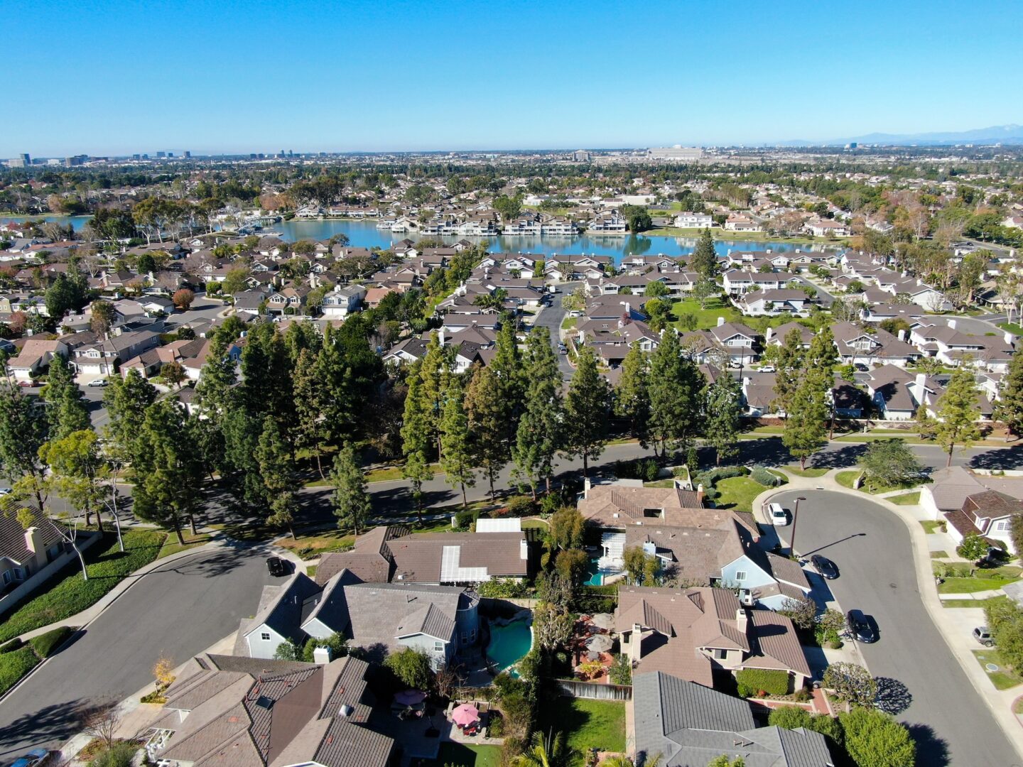 Aerial view of a suburban neighborhood with rows of houses, tree-lined streets, small backyards, and a large lake in the background under a clear blue sky. Aerial view of a suburban neighborhood with rows of houses, tree-lined streets, small backyards, and a large lake in the background under a clear blue sky.