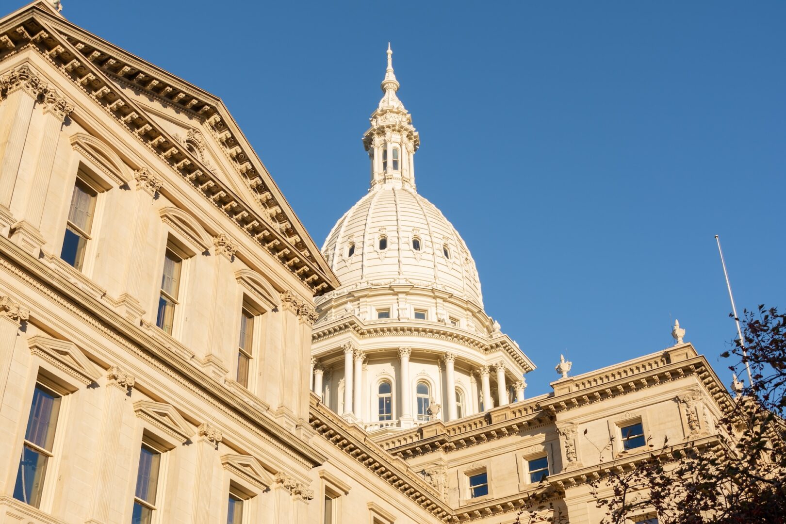 The image shows the Michigan State Capitol building in Lansing, featuring its white dome and ornate architectural details, set against a clear blue sky. The image shows the Michigan State Capitol building in Lansing, featuring its white dome and ornate architectural details, set against a clear blue sky.