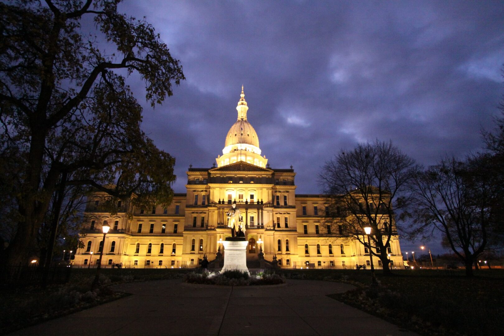 The Michigan State Capitol building illuminated at dusk, with a statue in front and trees framing the scene under a cloudy evening sky. The Michigan State Capitol building illuminated at dusk, with a statue in front and trees framing the scene under a cloudy evening sky.