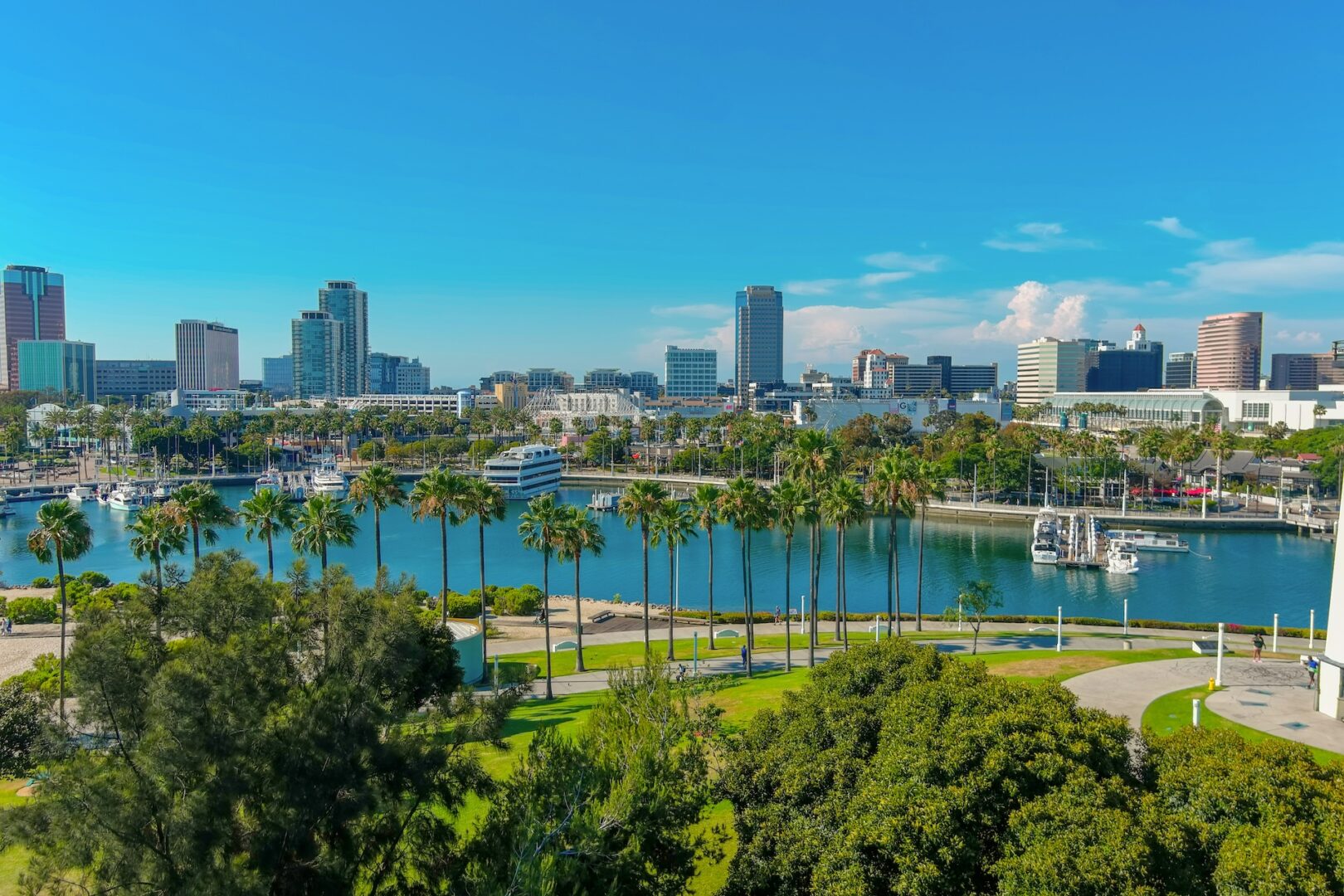 A view of a city skyline with tall buildings, palm trees, and a marina filled with boats along a calm blue waterway under a bright, clear sky. Green lawns and walking paths are visible in the foreground. A view of a city skyline with tall buildings, palm trees, and a marina filled with boats along a calm blue waterway under a bright, clear sky. Green lawns and walking paths are visible in the foreground.
