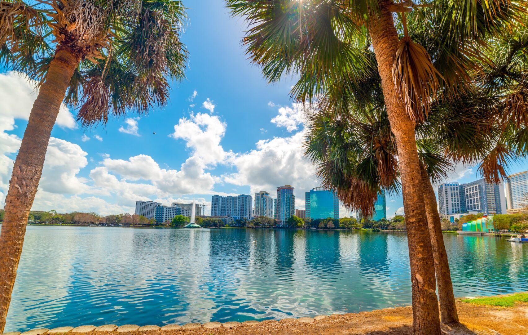 A scenic view of a city skyline with tall buildings reflected in a lake, framed by palm trees under a bright blue sky with scattered clouds. A scenic view of a city skyline with tall buildings reflected in a lake, framed by palm trees under a bright blue sky with scattered clouds.