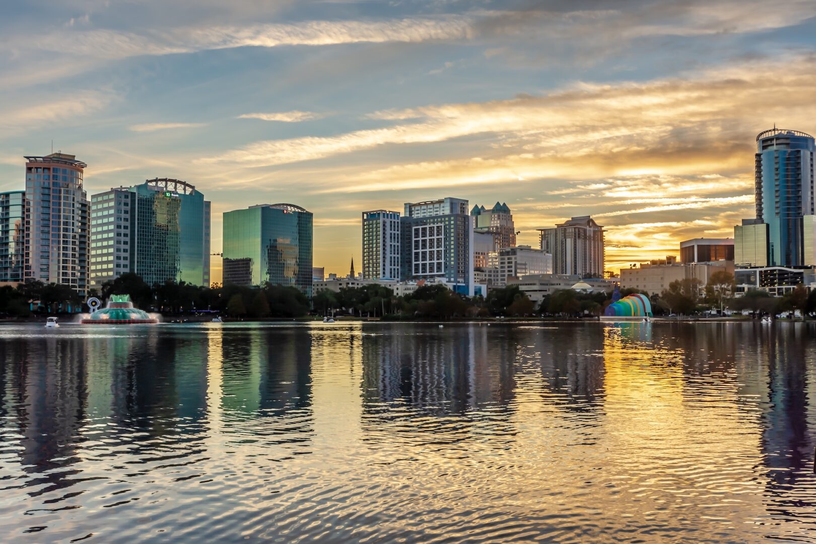 A city skyline with modern glass buildings is reflected in a calm lake at sunset. The sky is partly cloudy with golden and blue hues, and there are fountains and a colorful structure by the water’s edge. A city skyline with modern glass buildings is reflected in a calm lake at sunset. The sky is partly cloudy with golden and blue hues, and there are fountains and a colorful structure by the water’s edge.