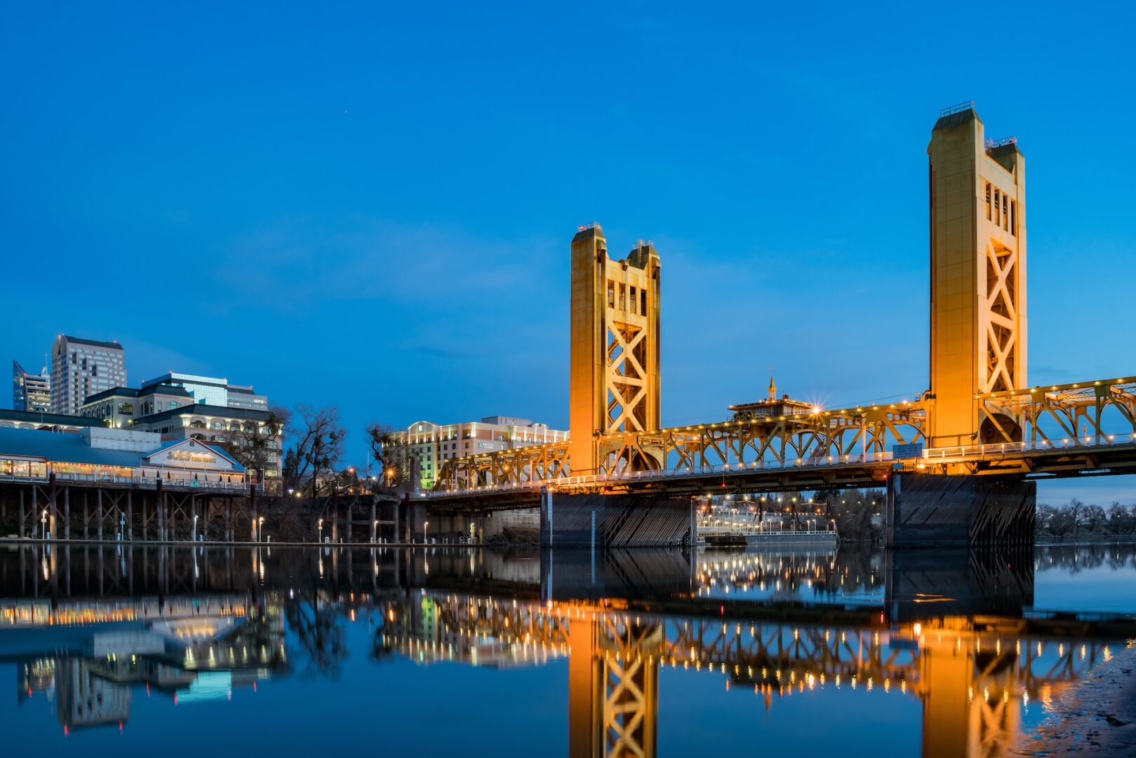 A golden-yellow tower bridge spans a calm river at dusk, reflecting the illuminated structure and nearby city buildings in the water under a clear blue sky. A golden-yellow tower bridge spans a calm river at dusk, reflecting the illuminated structure and nearby city buildings in the water under a clear blue sky.