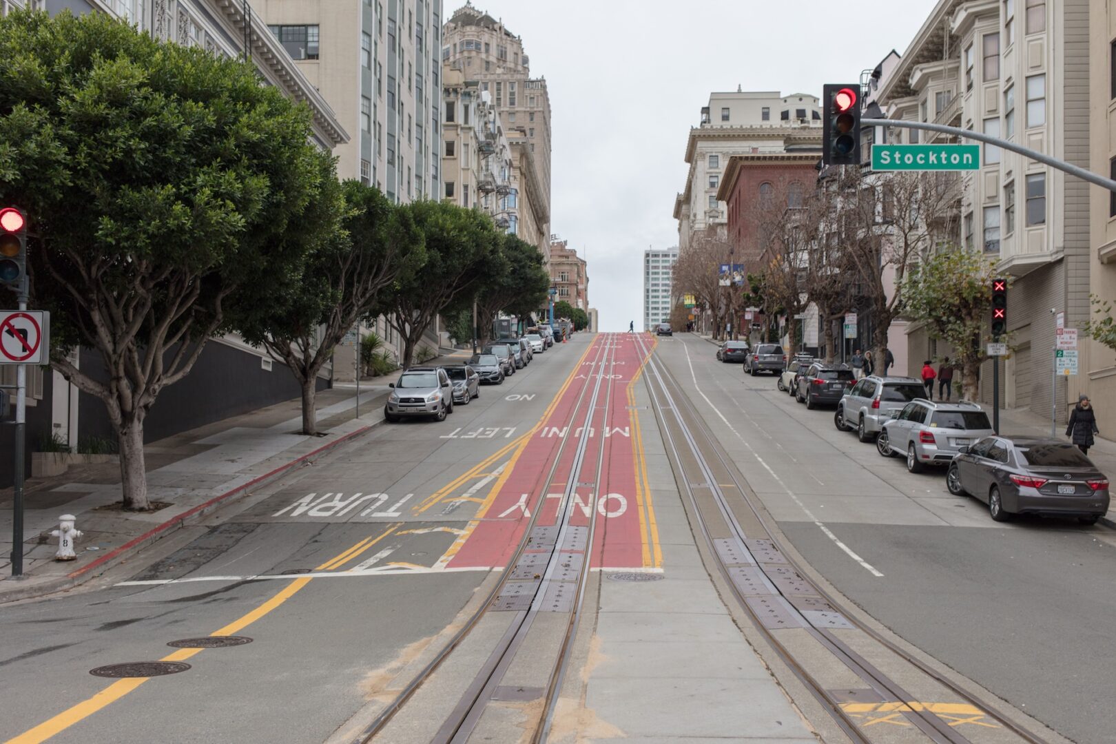 A steep, empty street in San Francisco with cable car tracks, lined with parked cars and tall buildings. A traffic light, street signs, and trees are visible along the sidewalk. The sky is overcast. A steep, empty street in San Francisco with cable car tracks, lined with parked cars and tall buildings. A traffic light, street signs, and trees are visible along the sidewalk. The sky is overcast.