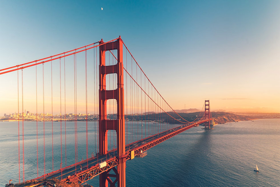 The Golden Gate Bridge spans across blue water at sunset, with a clear sky and the San Francisco skyline visible in the distance. The bridge's iconic red-orange towers and cables are bathed in warm light. The Golden Gate Bridge spans across blue water at sunset, with a clear sky and the San Francisco skyline visible in the distance. The bridge's iconic red-orange towers and cables are bathed in warm light.