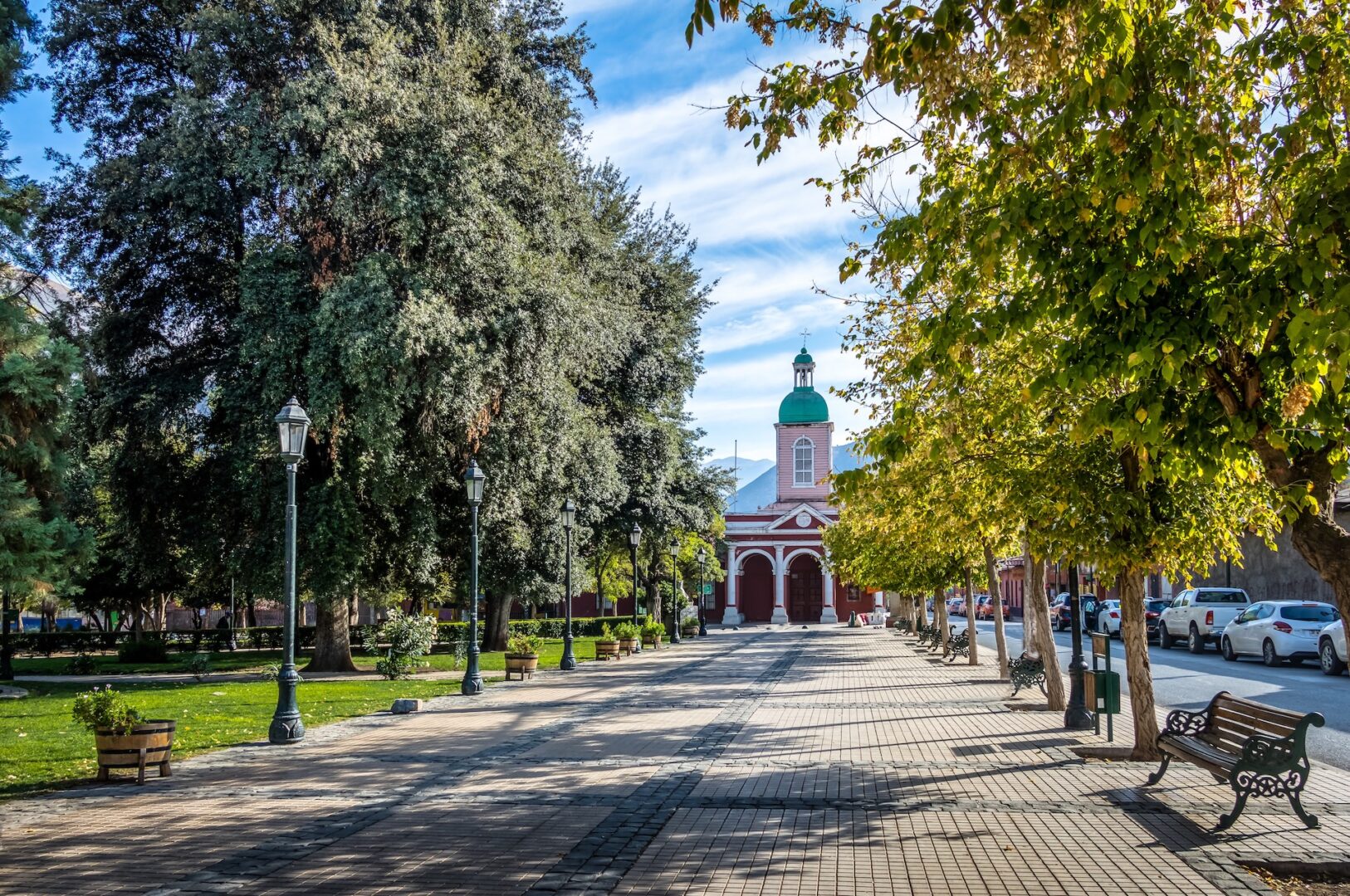 A tree-lined walkway with benches leads to a red church with a green dome under a partly cloudy sky; cars are parked along the street on the right. A tree-lined walkway with benches leads to a red church with a green dome under a partly cloudy sky; cars are parked along the street on the right.
