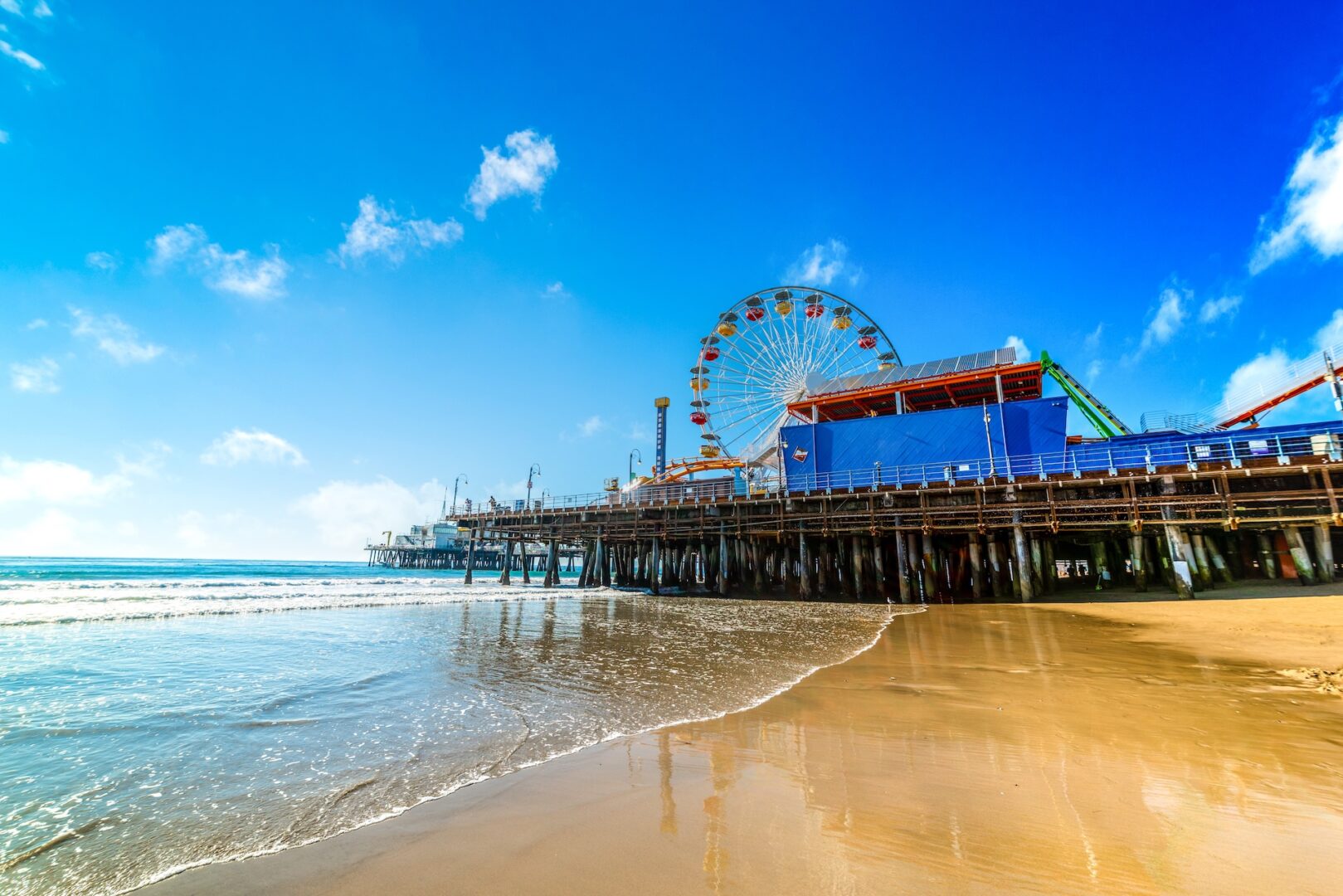 Santa Monica Pier with a colorful Ferris wheel and amusement rides sits above sandy beach and gentle ocean waves under a bright blue sky with scattered clouds. Santa Monica Pier with a colorful Ferris wheel and amusement rides sits above sandy beach and gentle ocean waves under a bright blue sky with scattered clouds.