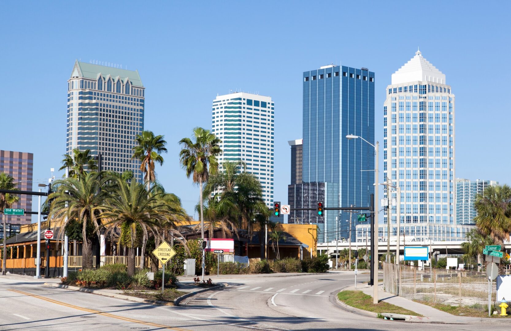 A cityscape featuring tall, modern skyscrapers and palm trees under a clear blue sky, with a road and traffic signals in the foreground. A cityscape featuring tall, modern skyscrapers and palm trees under a clear blue sky, with a road and traffic signals in the foreground.