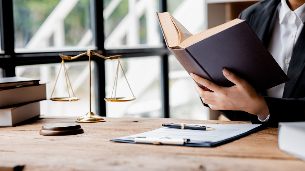 A person in business attire reads a book at a desk with legal documents, a pen, a gavel, and scales of justice, with large windows in the background. A person in business attire reads a book at a desk with legal documents, a pen, a gavel, and scales of justice, with large windows in the background.