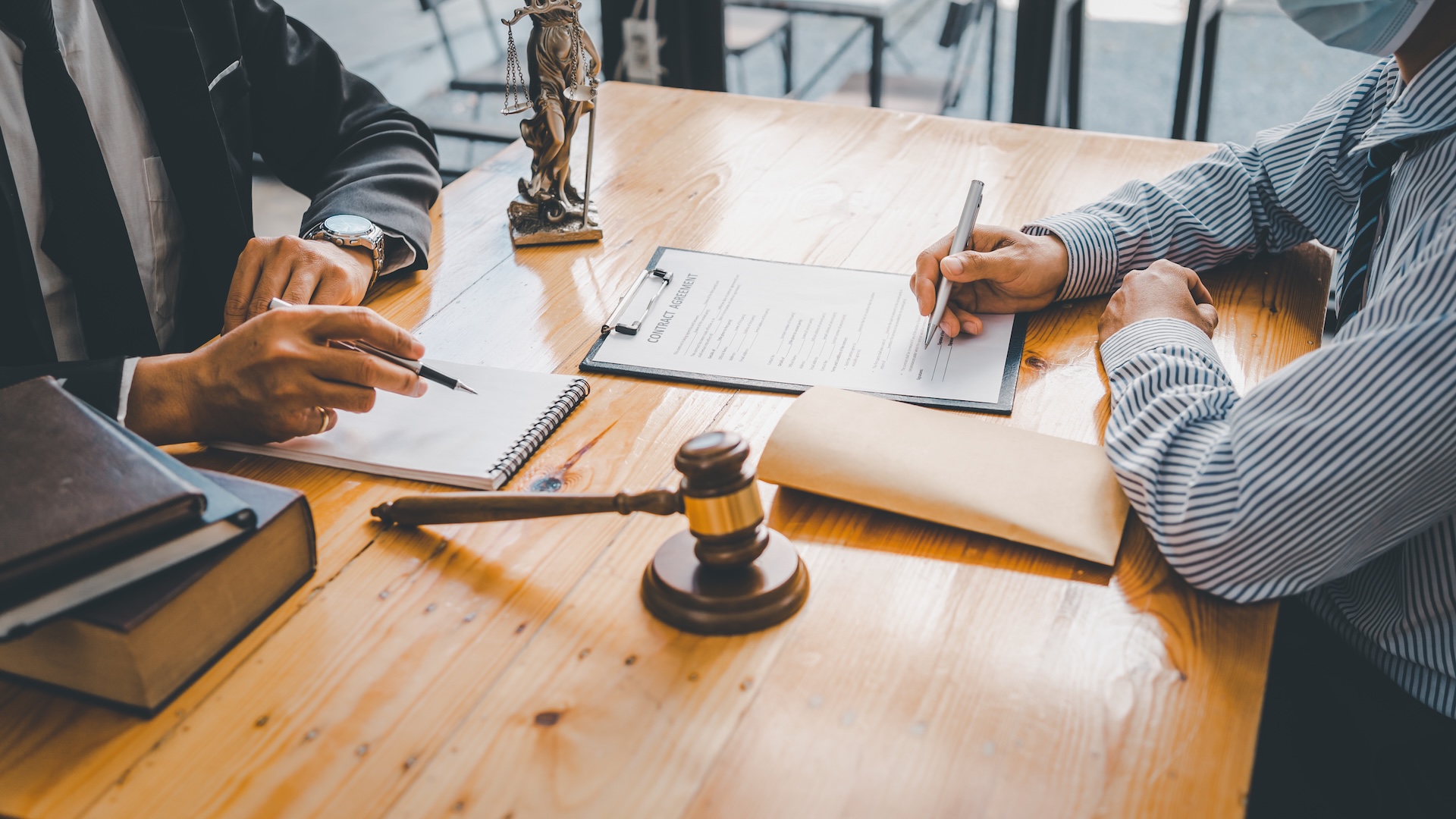 Two people sit at a wooden desk reviewing and signing documents, with legal books, a gavel, and a justice statue nearby, suggesting a legal or consultation setting. Two people sit at a wooden desk reviewing and signing documents, with legal books, a gavel, and a justice statue nearby, suggesting a legal or consultation setting.