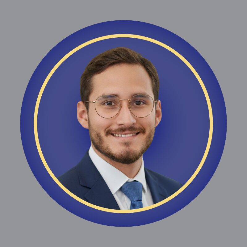 A man with short brown hair, a trimmed beard, and glasses, wearing a dark suit, white shirt, and blue tie, smiles at the camera. He is framed by a circular blue and yellow border on a gray background. A man with short brown hair, a trimmed beard, and glasses, wearing a dark suit, white shirt, and blue tie, smiles at the camera. He is framed by a circular blue and yellow border on a gray background.